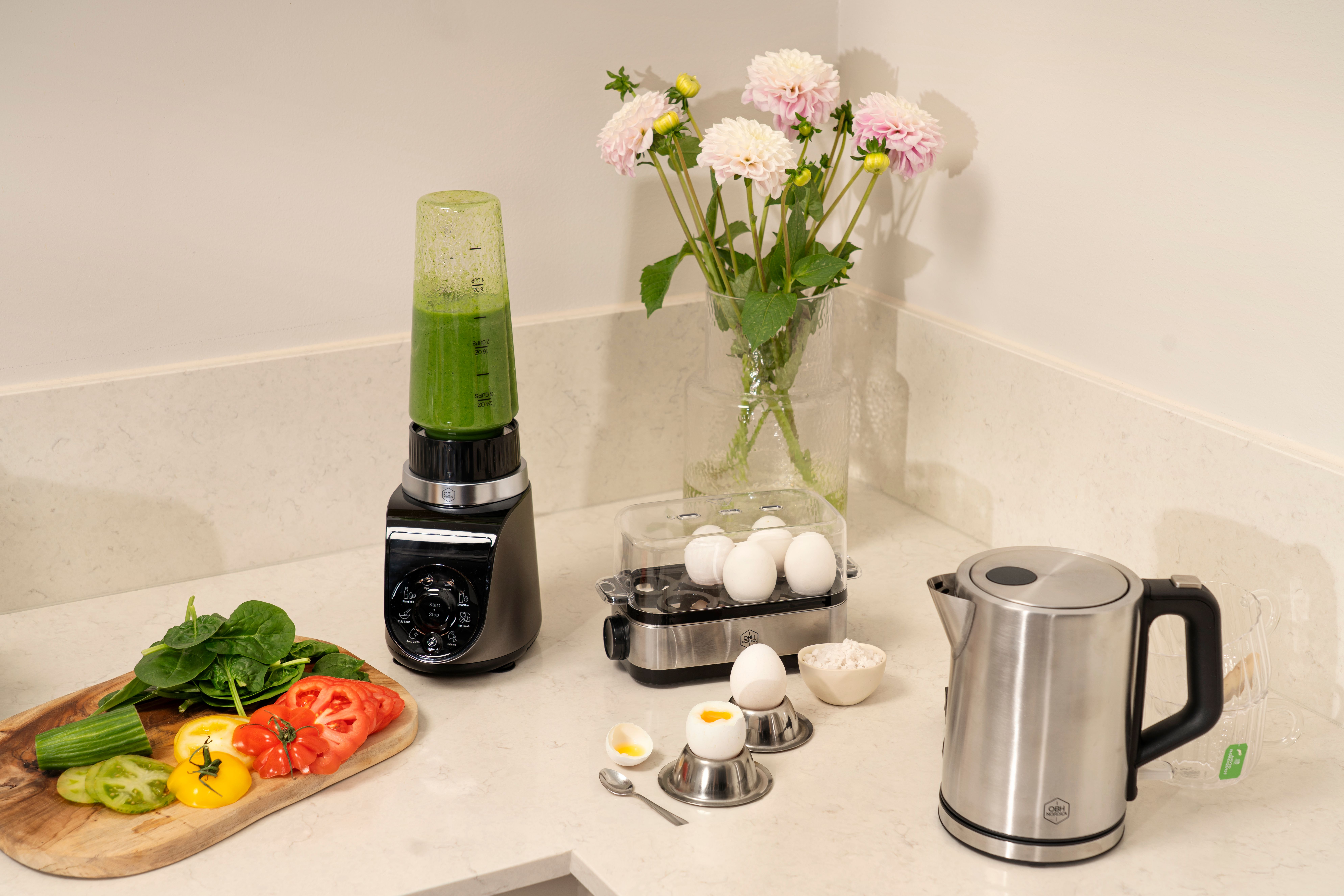 A blender with green smoothie, sliced vegetables, egg cooker, soft-boiled egg, electric kettle, and flowers on a kitchen counter.