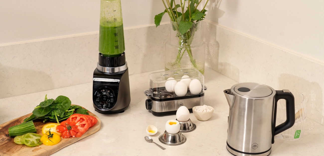 A blender with green smoothie, sliced vegetables, egg cooker, soft-boiled egg, electric kettle, and flowers on a kitchen counter.
