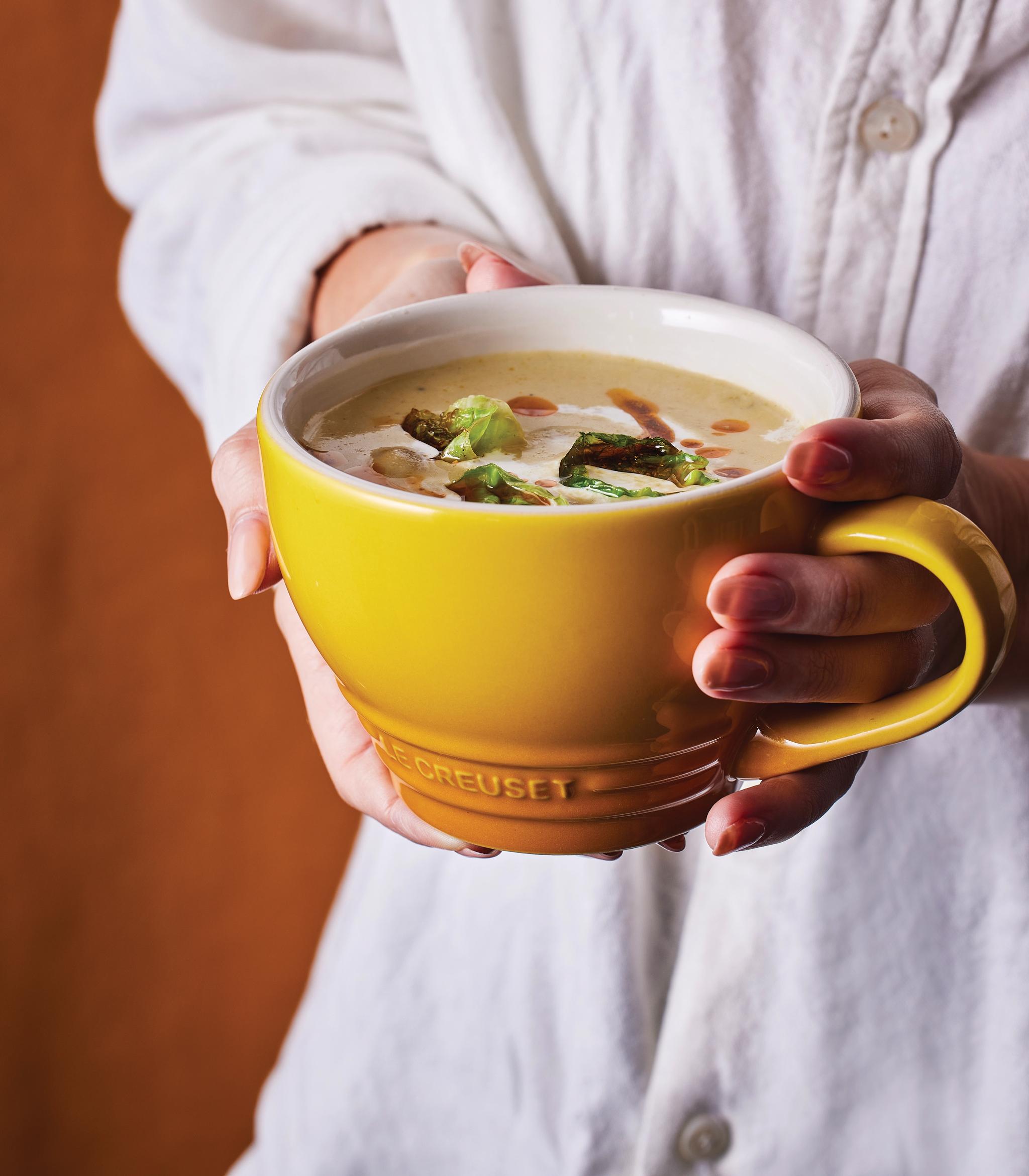 En person i vit skjorta håller en gul mugg krämig soppa garnerad med rostade gröna grönsaker.