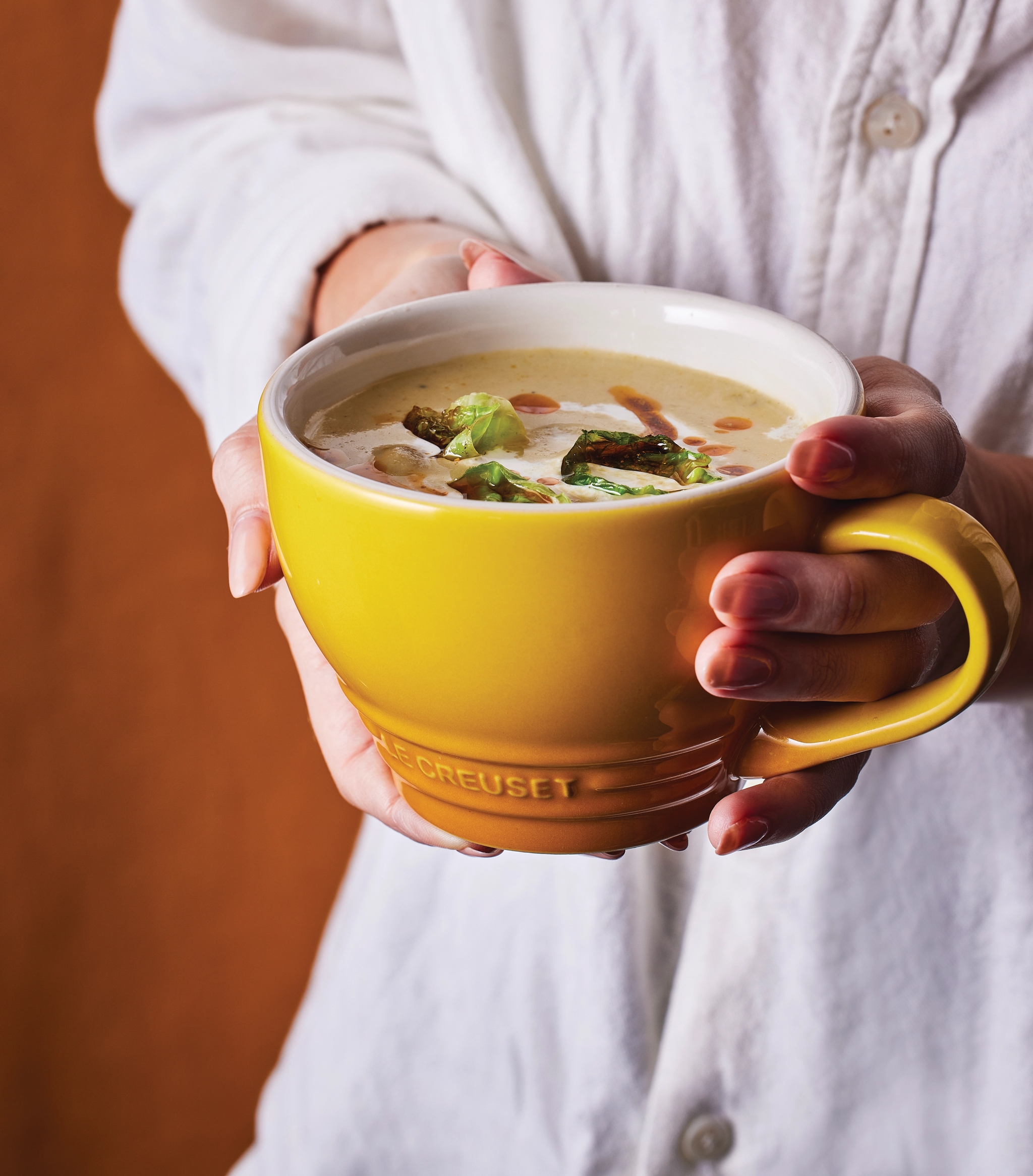 En person i vit skjorta håller en gul mugg krämig soppa garnerad med rostade gröna grönsaker.