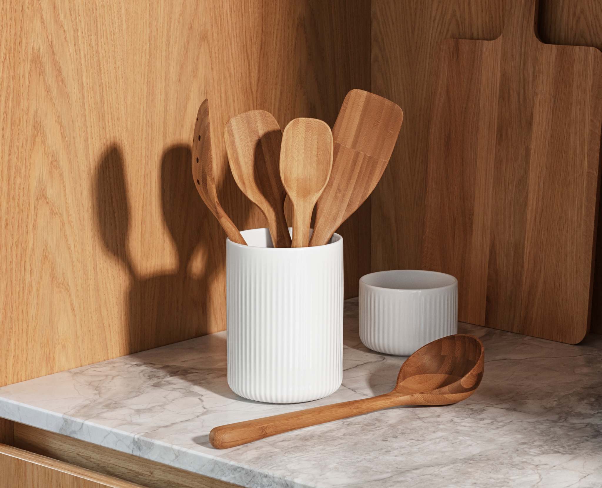 Valkoisissa uurretuissa keraamisissa astioissa on puisia keittiövälineitä ja artisokkaa marmorisella tiskillä puukaappeja vasten.