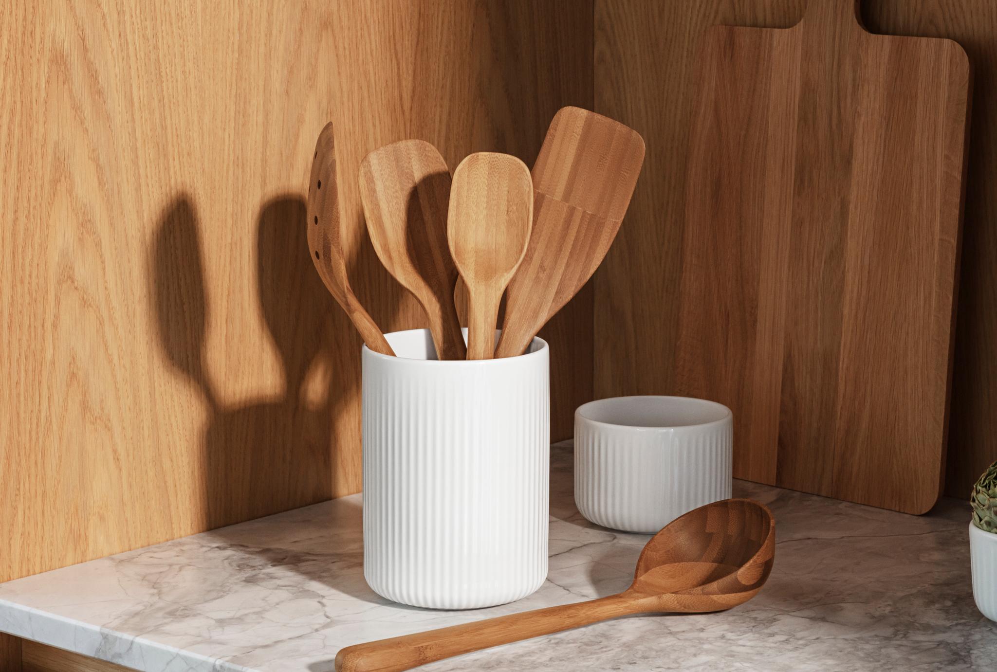 Valkoisissa uurretuissa keraamisissa astioissa on puisia keittiövälineitä ja artisokkaa marmorisella tiskillä puukaappeja vasten.