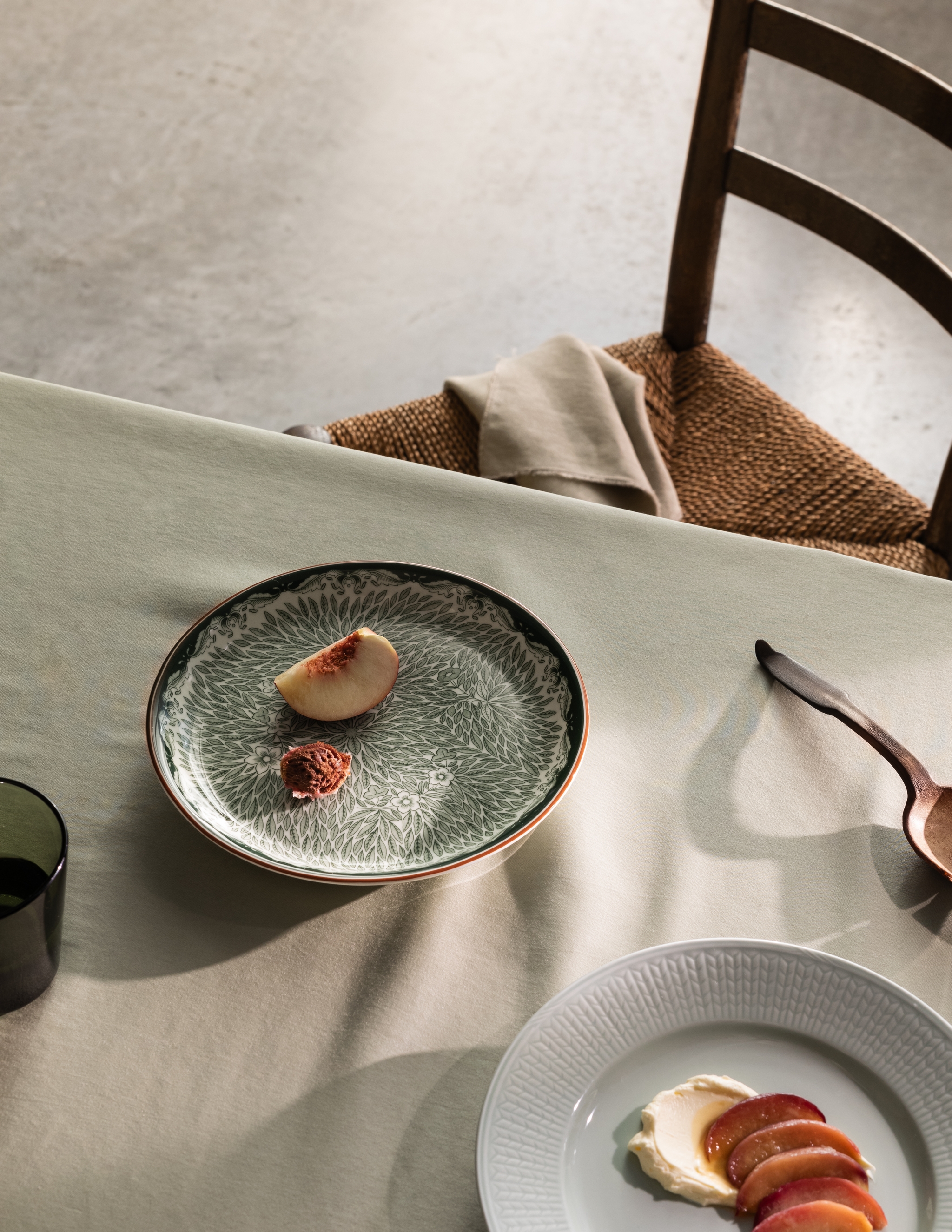 Matplats med beige duk, rustik trästol och enkel dessertservering på mönstrat grönt fat och vit tallrik, tillsammans med träsked och linneservett i naturligt ljus.