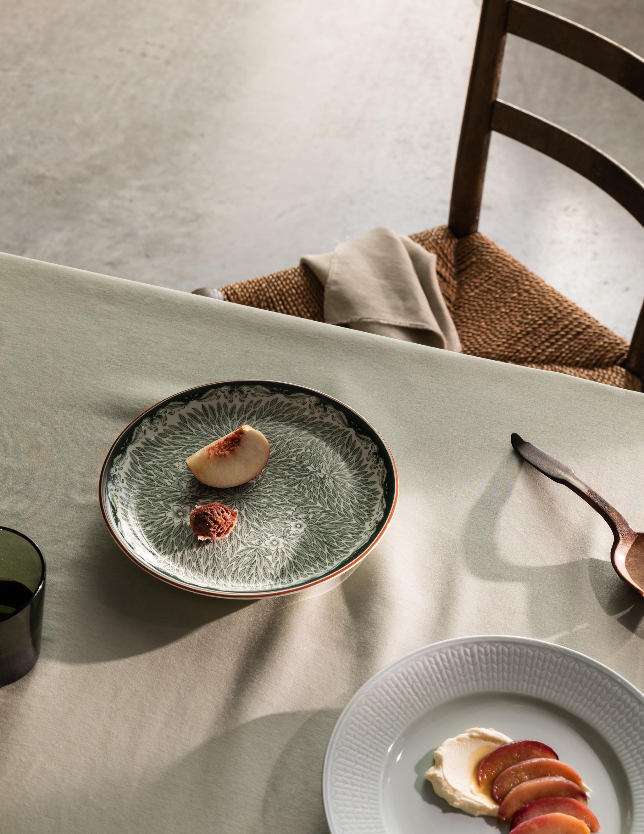 Matplats med beige duk, rustik trästol och enkel dessertservering på mönstrat grönt fat och vit tallrik, tillsammans med träsked och linneservett i naturligt ljus.