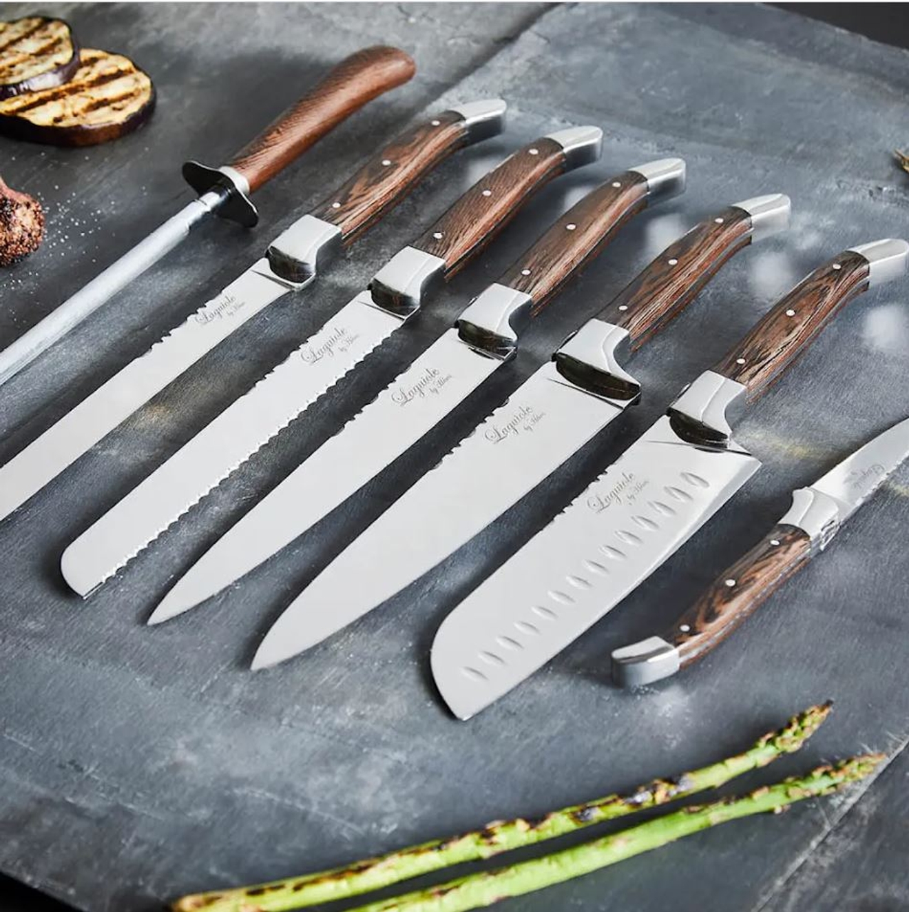 Exklusivt set med köksknivar med trähandtag upplagda på en mörk skärbräda bredvid grillade grönsaker.