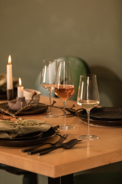 Ett matbord dukat med tända ljus, vinglas med rosé- och vitt vin, mörka tallrikar och naturlig grendekor.