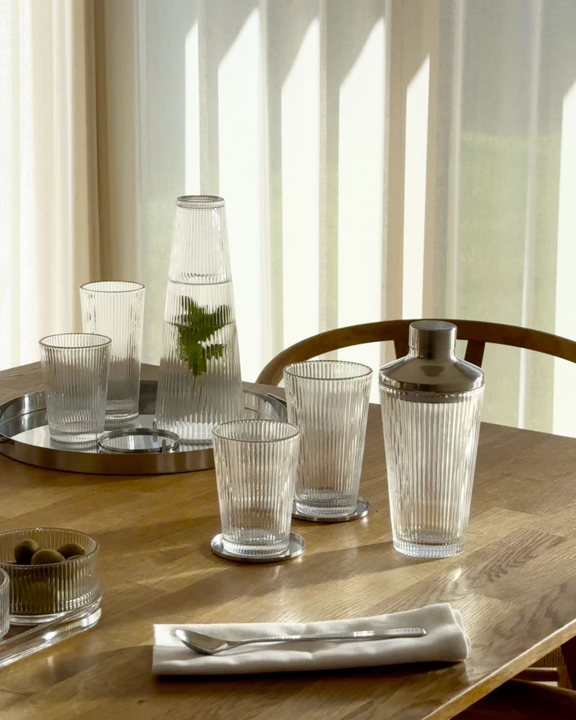 Elegant dukning med räfflade glas, karaff och shaker i klart glas på ett solbelyst träbord.