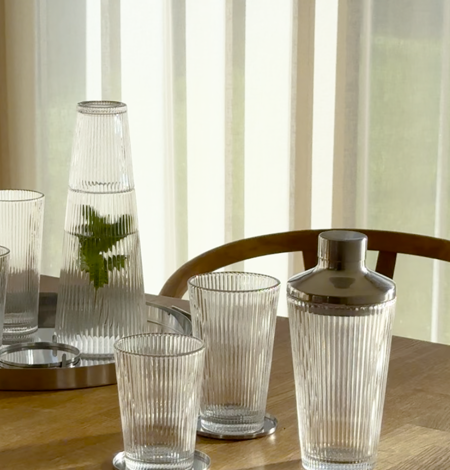 Elegant dukning med räfflade glas, karaff och shaker i klart glas på ett solbelyst träbord.