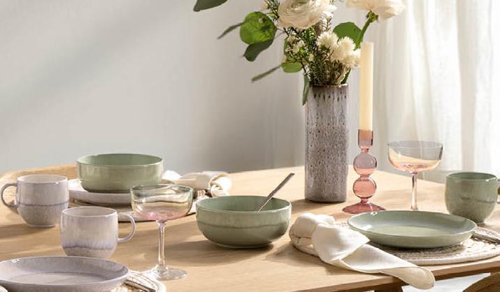 Vaaleapuinen ruokapöytä on katettu pastellivärisillä astioilla, kukka-asetelmalla ja kynttilöillä, ja sitä ympäröivät puiset tuolit valoisassa huoneessa.