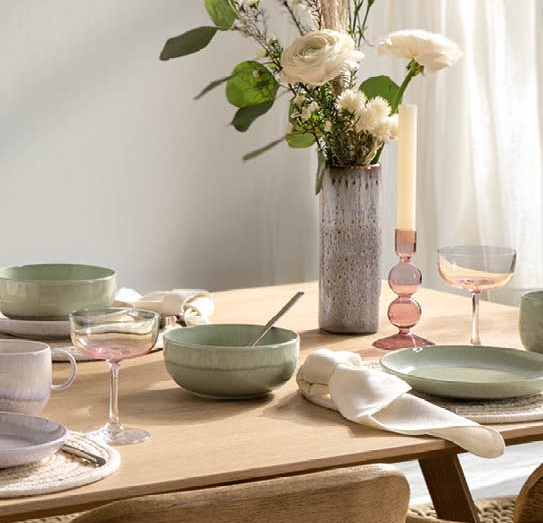 Vaaleapuinen ruokapöytä on katettu pastellivärisillä astioilla, kukka-asetelmalla ja kynttilöillä, ja sitä ympäröivät puiset tuolit valoisassa huoneessa.