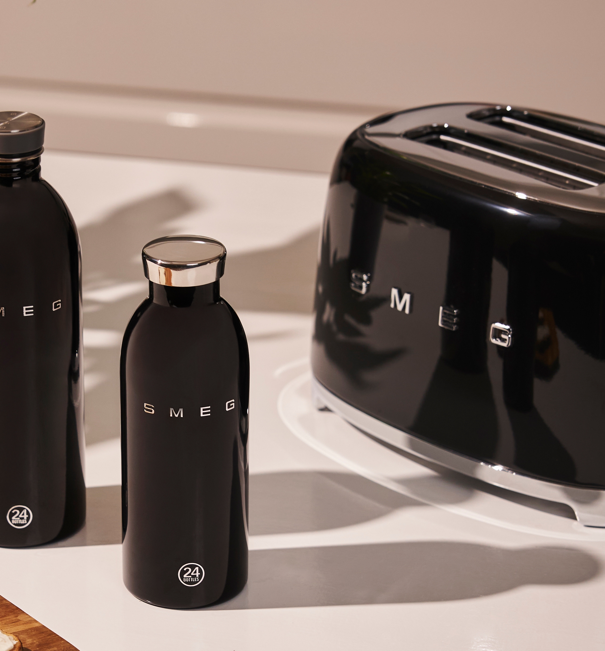 Musta Smeg-leivänpaahdin, kaksi mustaa Smeg-vesipulloa ja paahtoleipää valkoisella levitteellä puulaudalla valkoisella tiskillä.