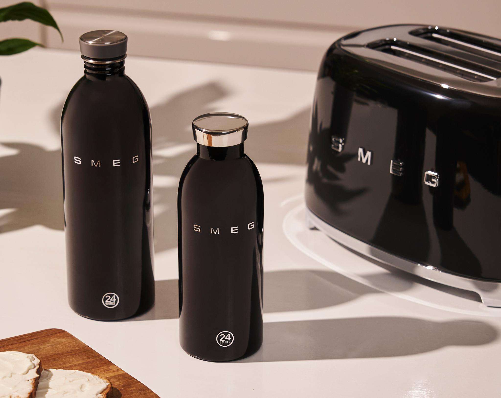 Musta Smeg-leivänpaahdin, kaksi mustaa Smeg-vesipulloa ja paahtoleipää valkoisella levitteellä puulaudalla valkoisella tiskillä.