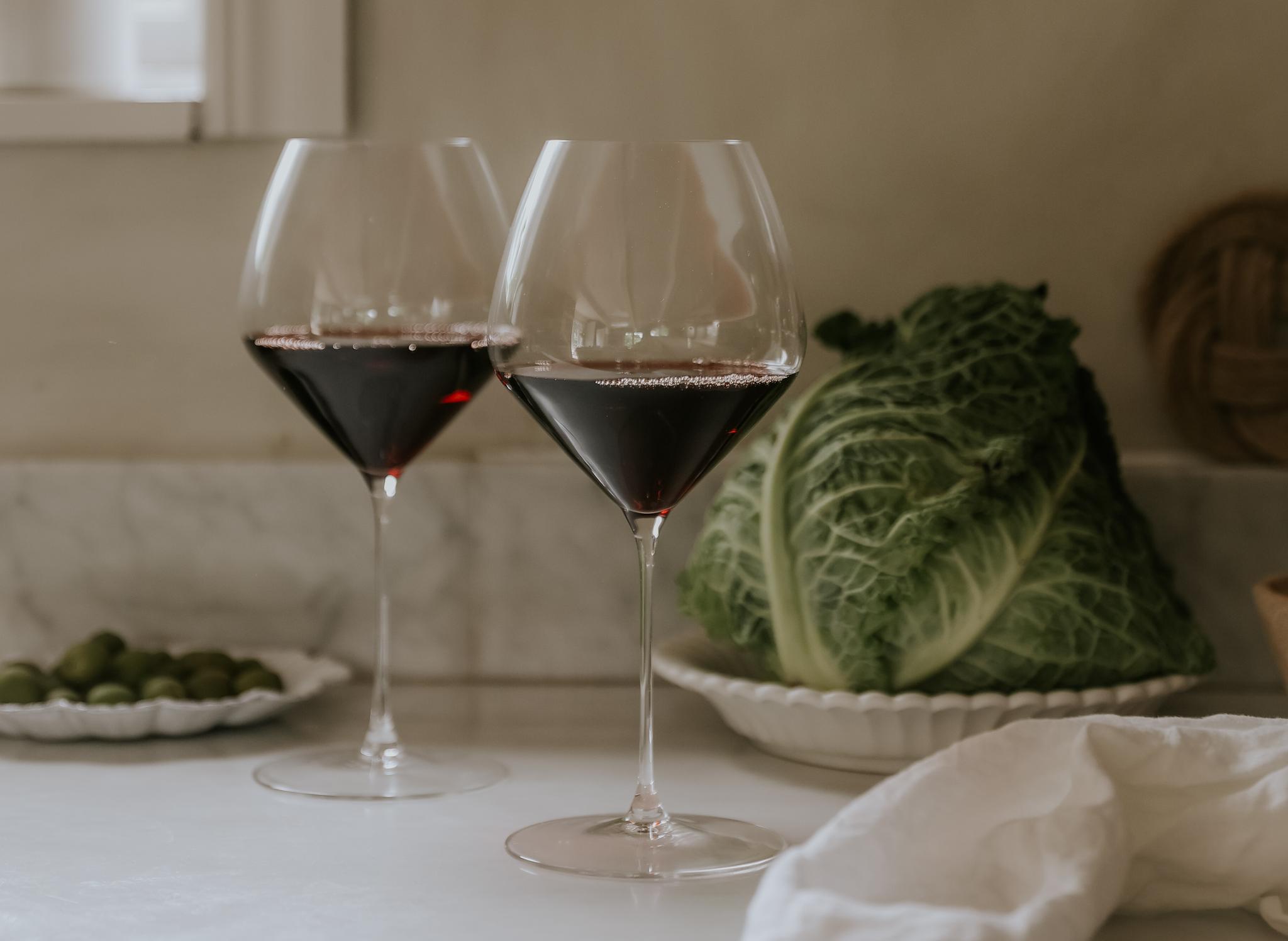 Två glas rött vin på en köksbänk i vit marmor, bredvid ett kålhuvud, gröna oliver och en vit linneduk.