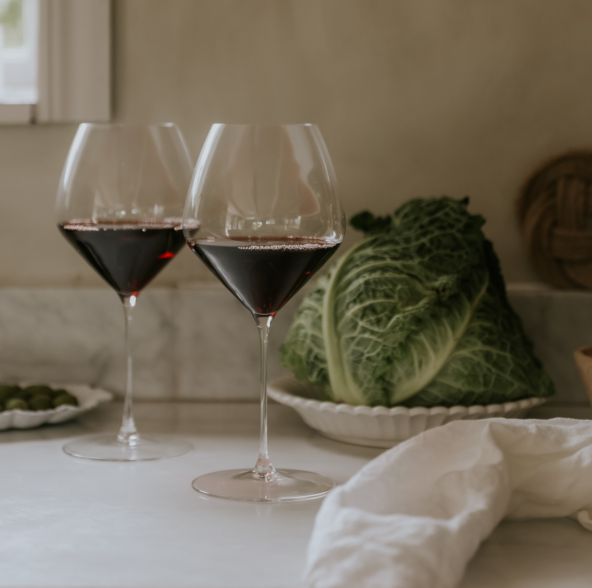Två glas rött vin på en köksbänk i vit marmor, bredvid ett kålhuvud, gröna oliver och en vit linneduk.