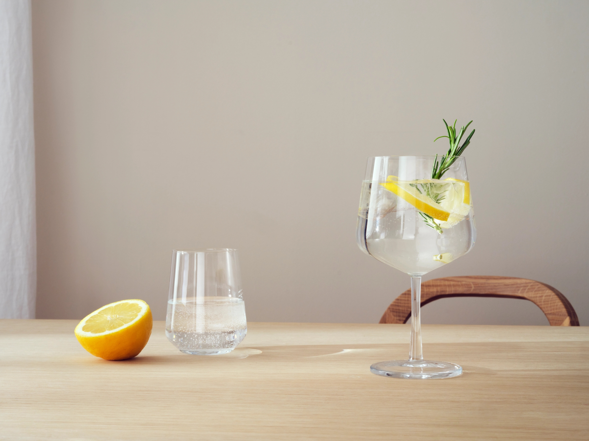 Et glass med stilk med en drink garnert med sitron og rosmarin, et mindre glass vann og en oppskåret sitron på et lyst trebord.