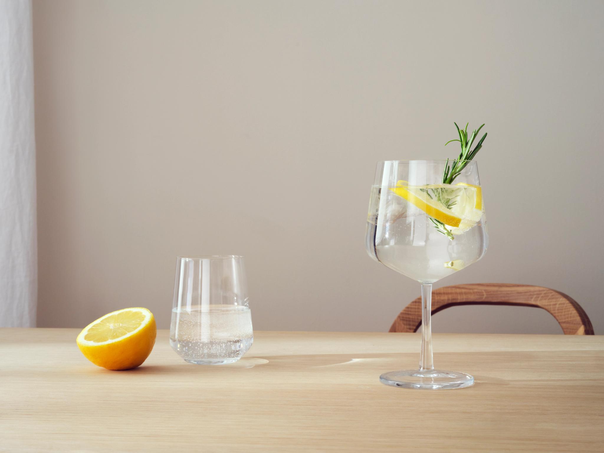Et glass med stilk med en drink garnert med sitron og rosmarin, et mindre glass vann og en oppskåret sitron på et lyst trebord.