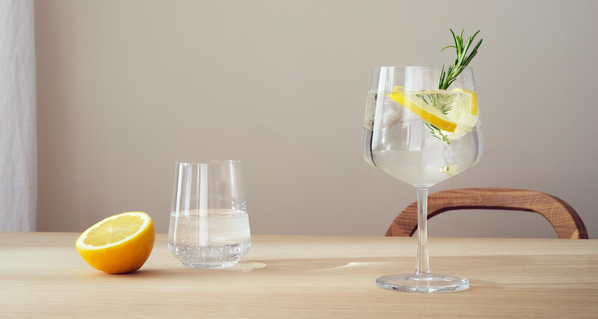Vaalealla puupöydällä on puolitettu sitruuna, lasillinen kivennäisvettä ja varrellinen vesilasi, koristeltuna sitruunalla ja yrteillä.