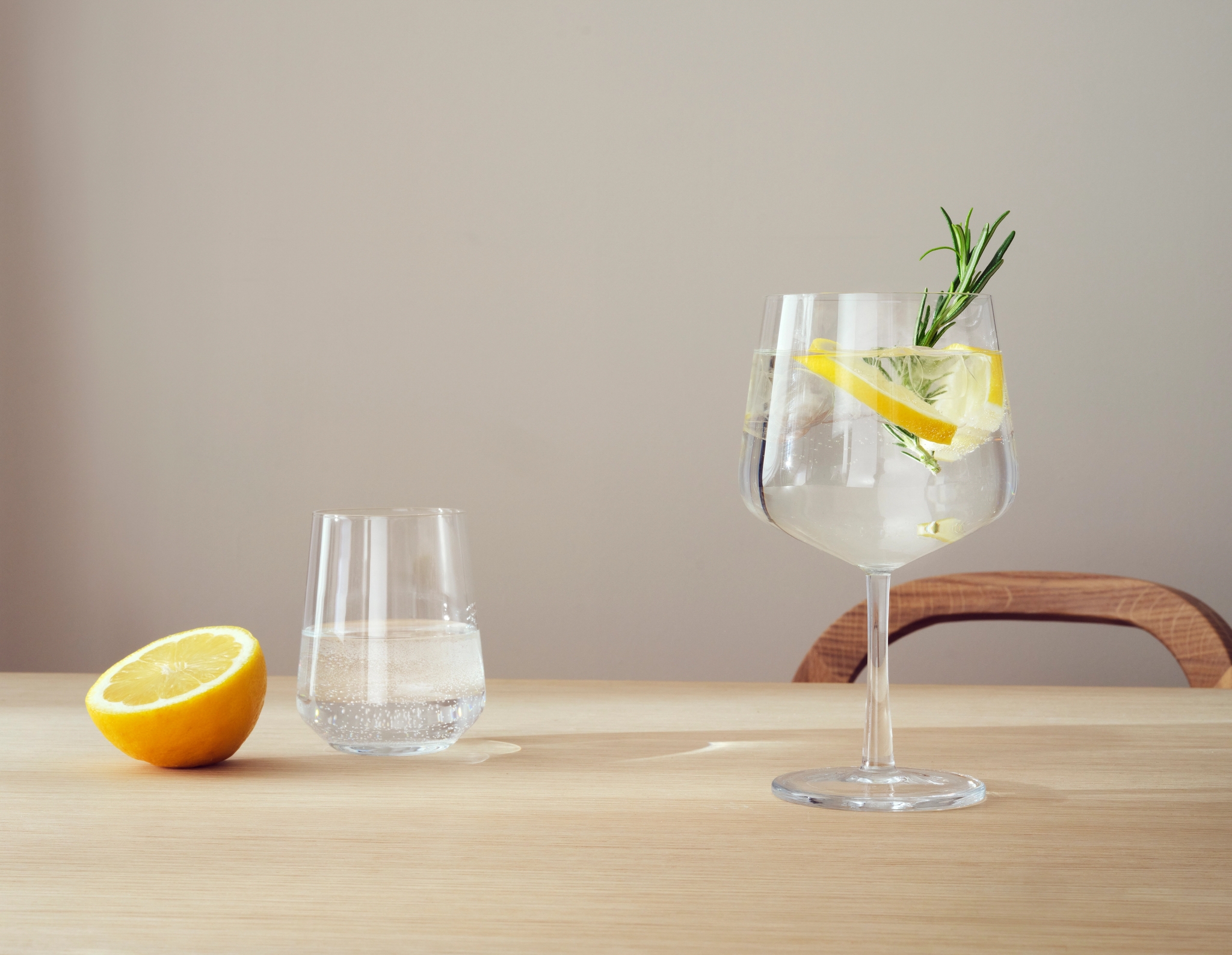Ett glas med klar dryck på stjälk garnerat med citron och rosmarin, ett glas vatten och en halv citron på ett träbord.