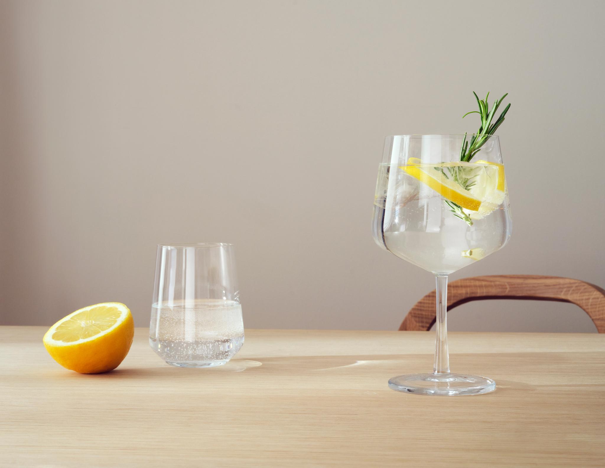 Ett glas med klar dryck på stjälk garnerat med citron och rosmarin, ett glas vatten och en halv citron på ett träbord.