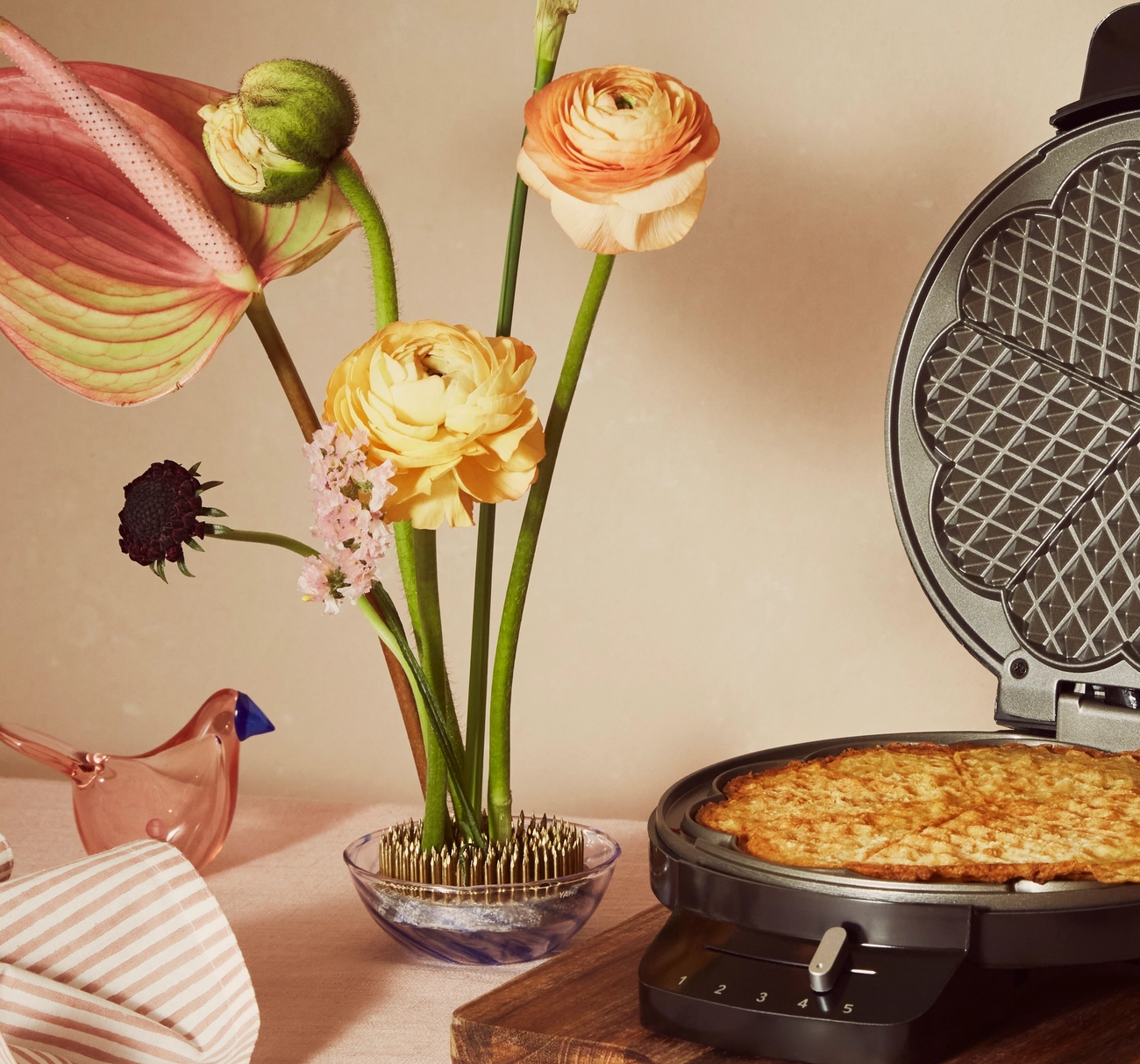 Kukkasiili jossa kukka-asetelma pöydällä Iittala lasilinnun ja vohveliraudan kera