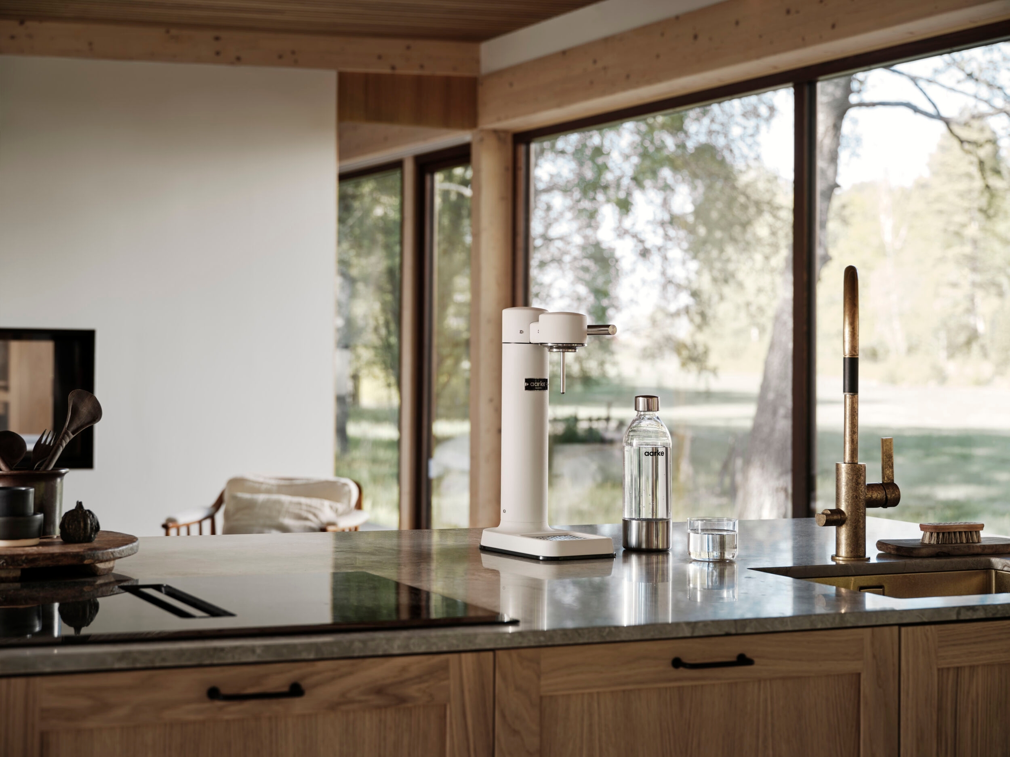 Modernt kök med kolsyremaskin, glasflaska och mässingskran på en köksö framför stora fönster mot grönskande natur.