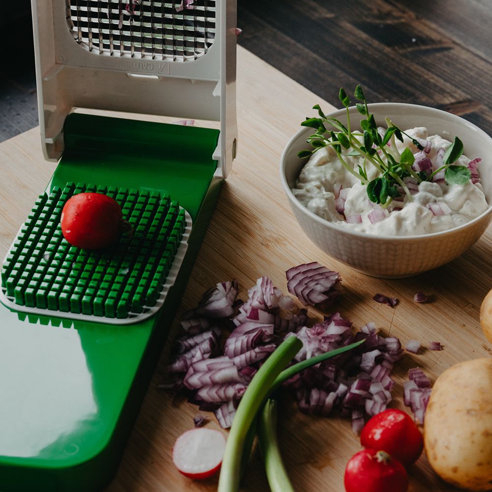 En grønnsakshakker med en reddik på risten, ved siden av en bolle med dip, hakket rødløk og andre grønnsaker på et skjærebrett i tre.