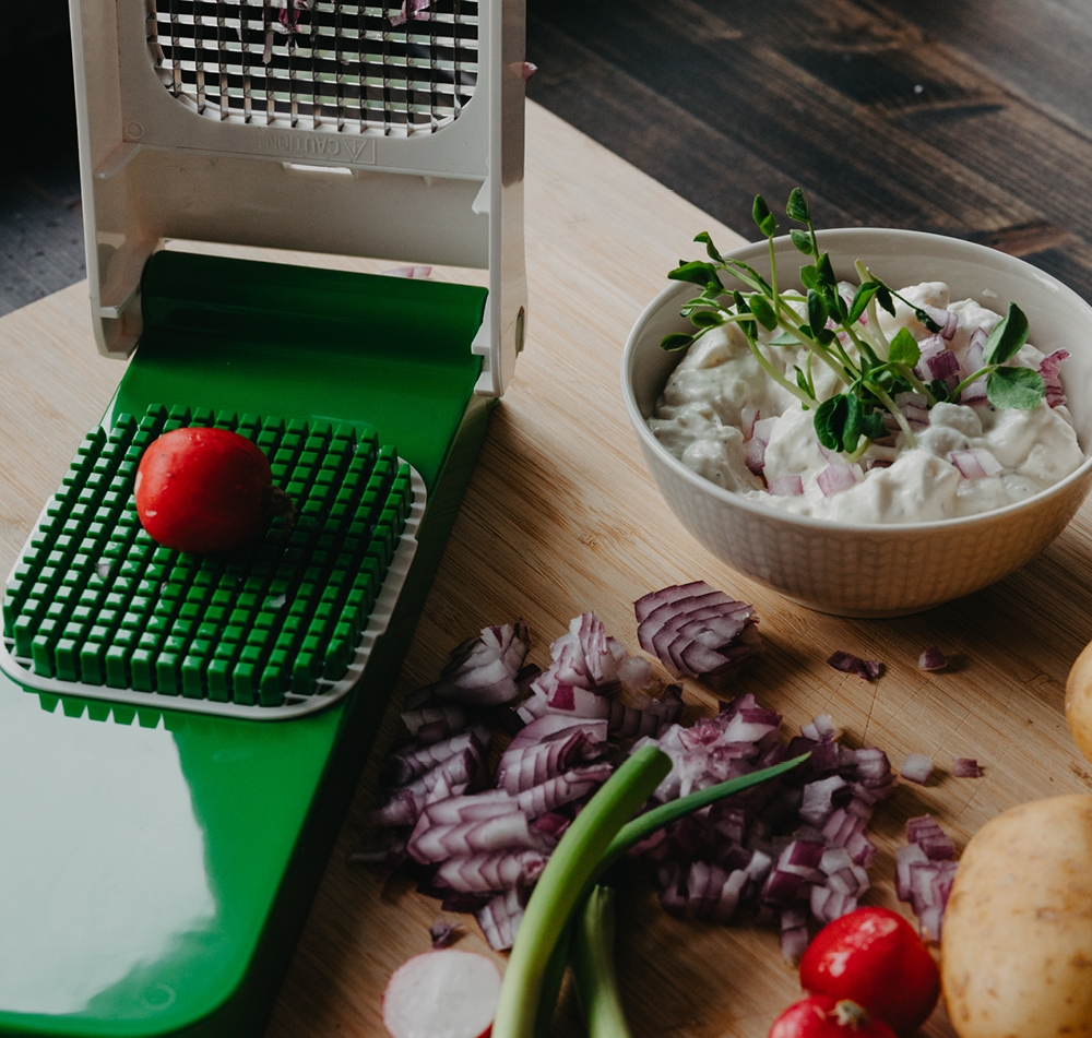 En grönsakshackare med en rädisa på sitt rutnät, bredvid en skål med dippsås, hackad rödlök och andra grönsaker på en träskärbräda.