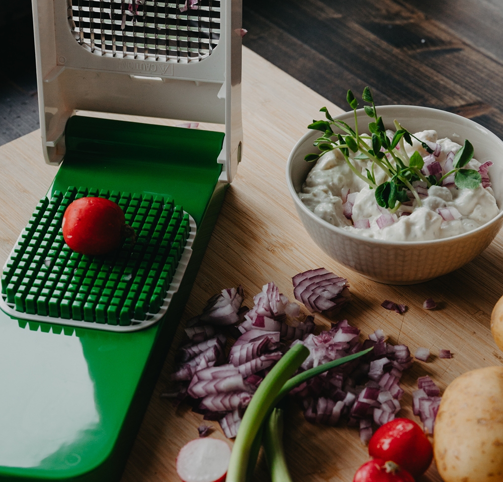 Vihannesleikkuri, jossa on retiisi ritilän päällä, vieressä kulho dippiä, pilkottua punasipulia ja muita vihanneksia puisella leikkuulaudalla.