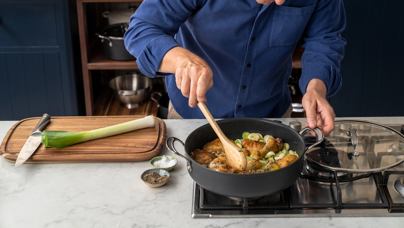 A man in a blue shirt stirring chicken and leeks in a pan on a stovetop.