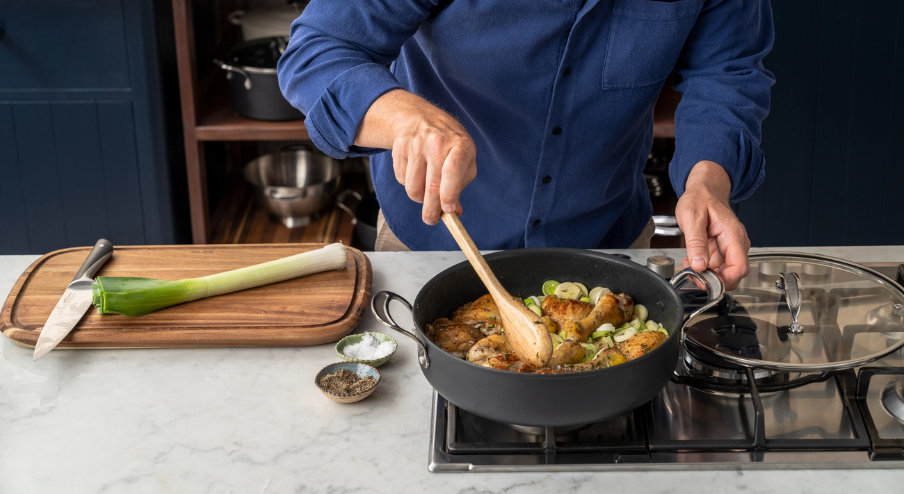 A man in a blue shirt stirs chicken and leeks in a pan on a stove in a kitchen.