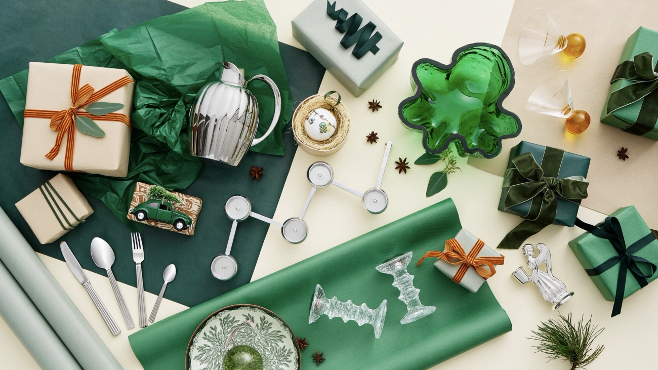 A festive flat lay featuring green wrapped gifts, silver homeware, green tissue paper, and holiday decorations on a two-toned background.