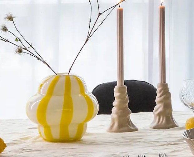 Keltainen maljakko valkoisilla raidoilla keittiön pöydällä kynttilänjalkojen ja kulhon kera