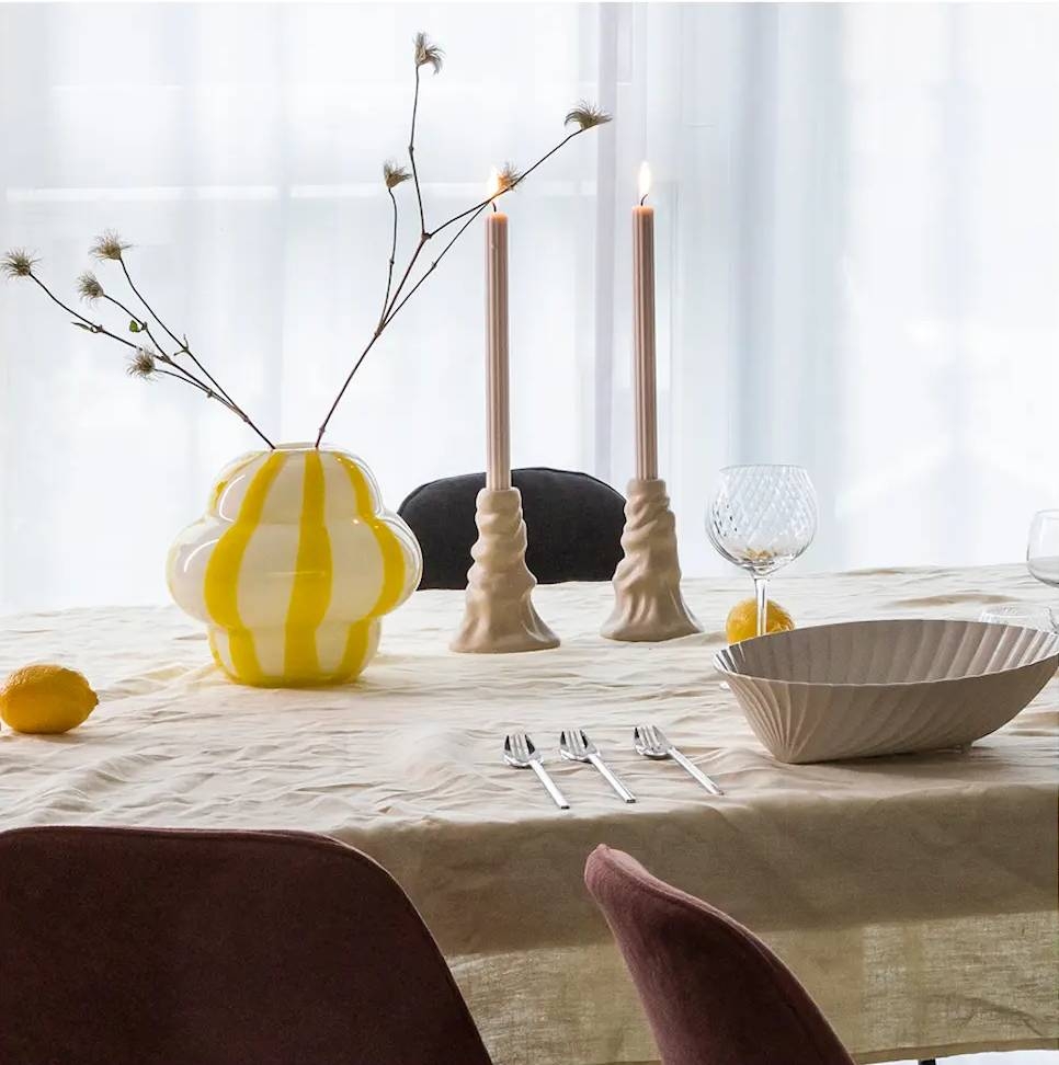 Keltainen maljakko valkoisilla raidoilla keittiön pöydällä kynttilänjalkojen ja kulhon kera