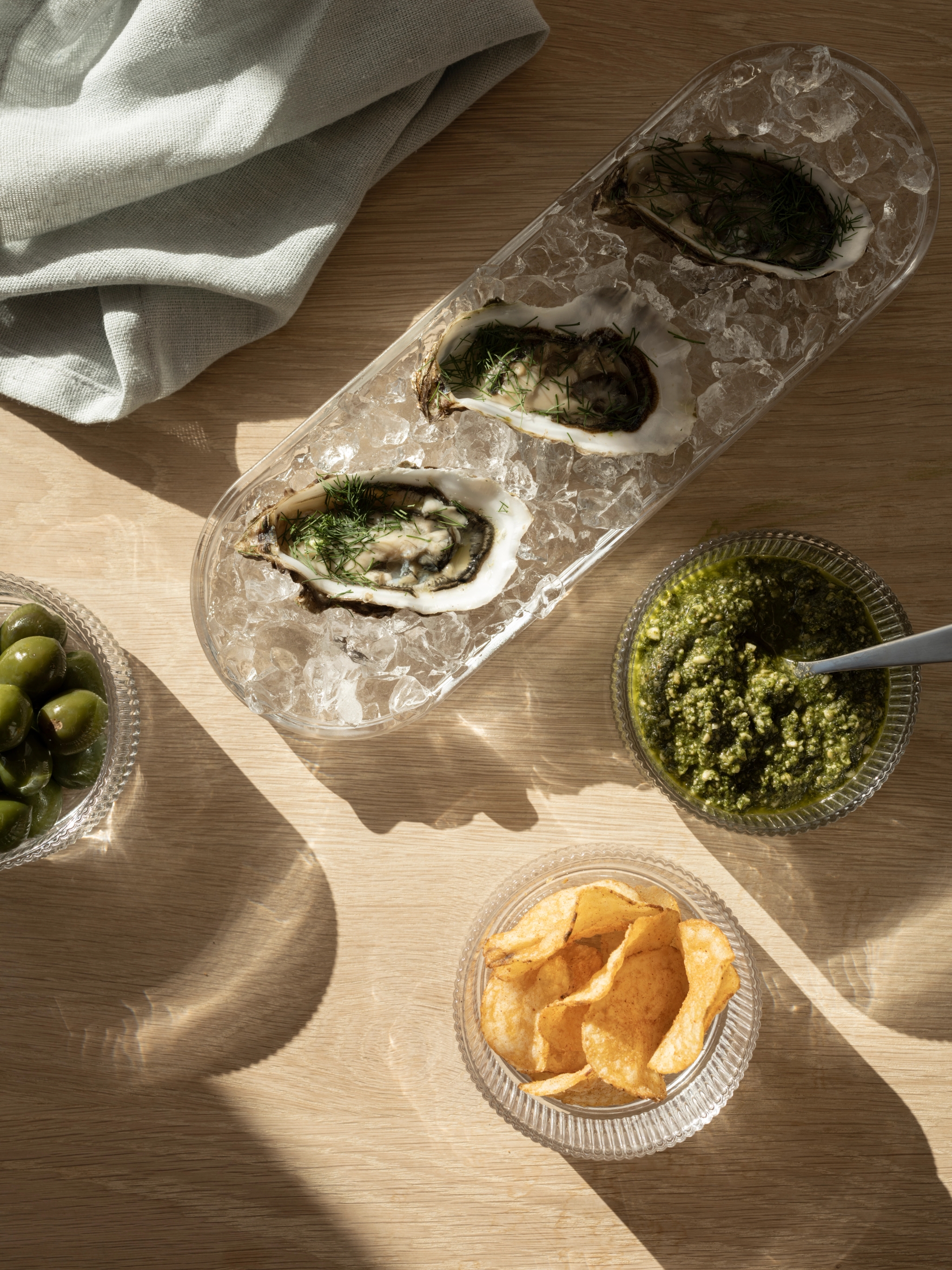 Stillben med ostron på is, gröna oliver, chips och en skål med grön pesto på ett ljust träbord i solljus.