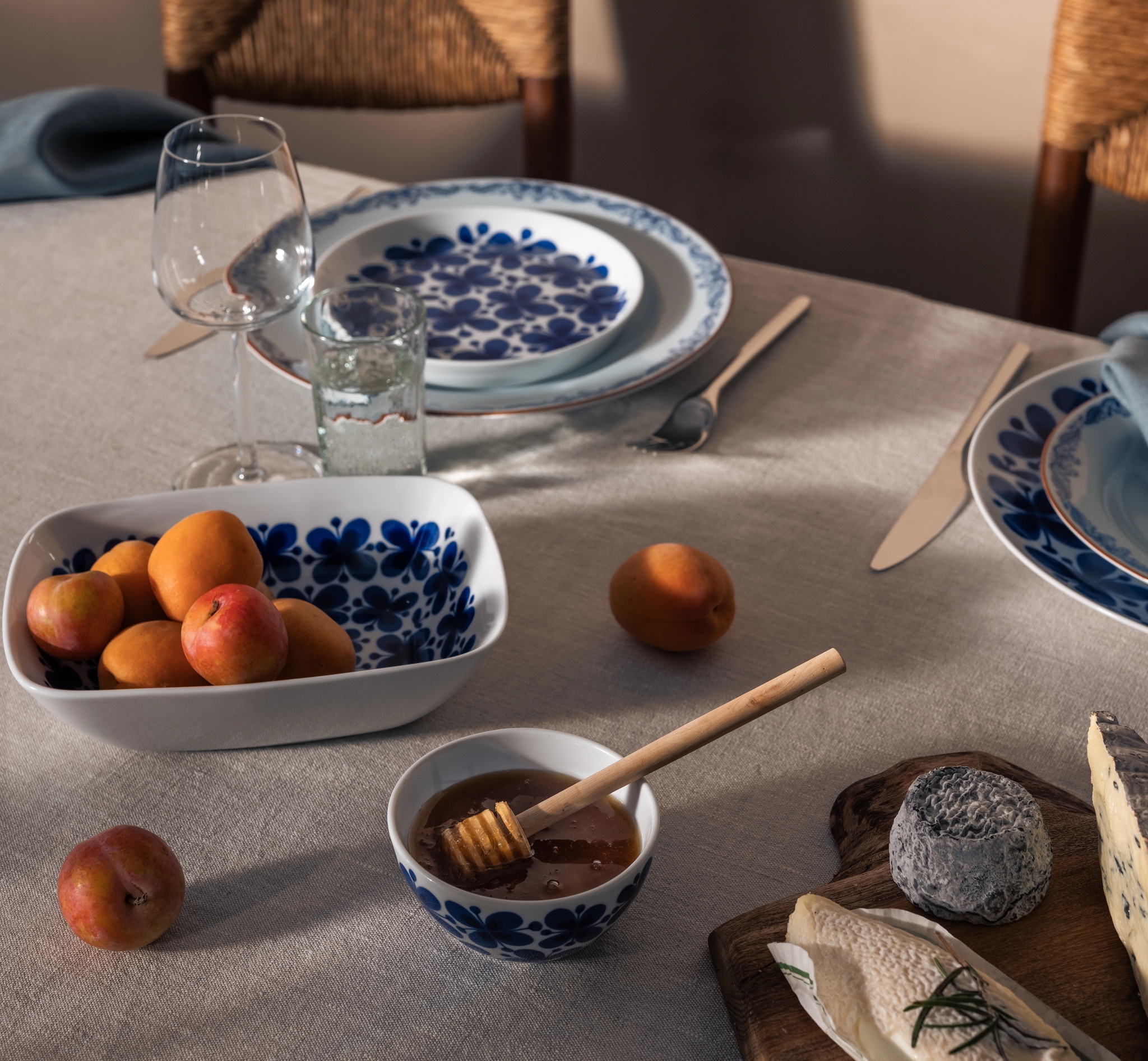 Ett bord dukat med blåvita mönstrade fat, en skål med aprikoser och plommon, honung och en ostbricka, upplyst av solljus.