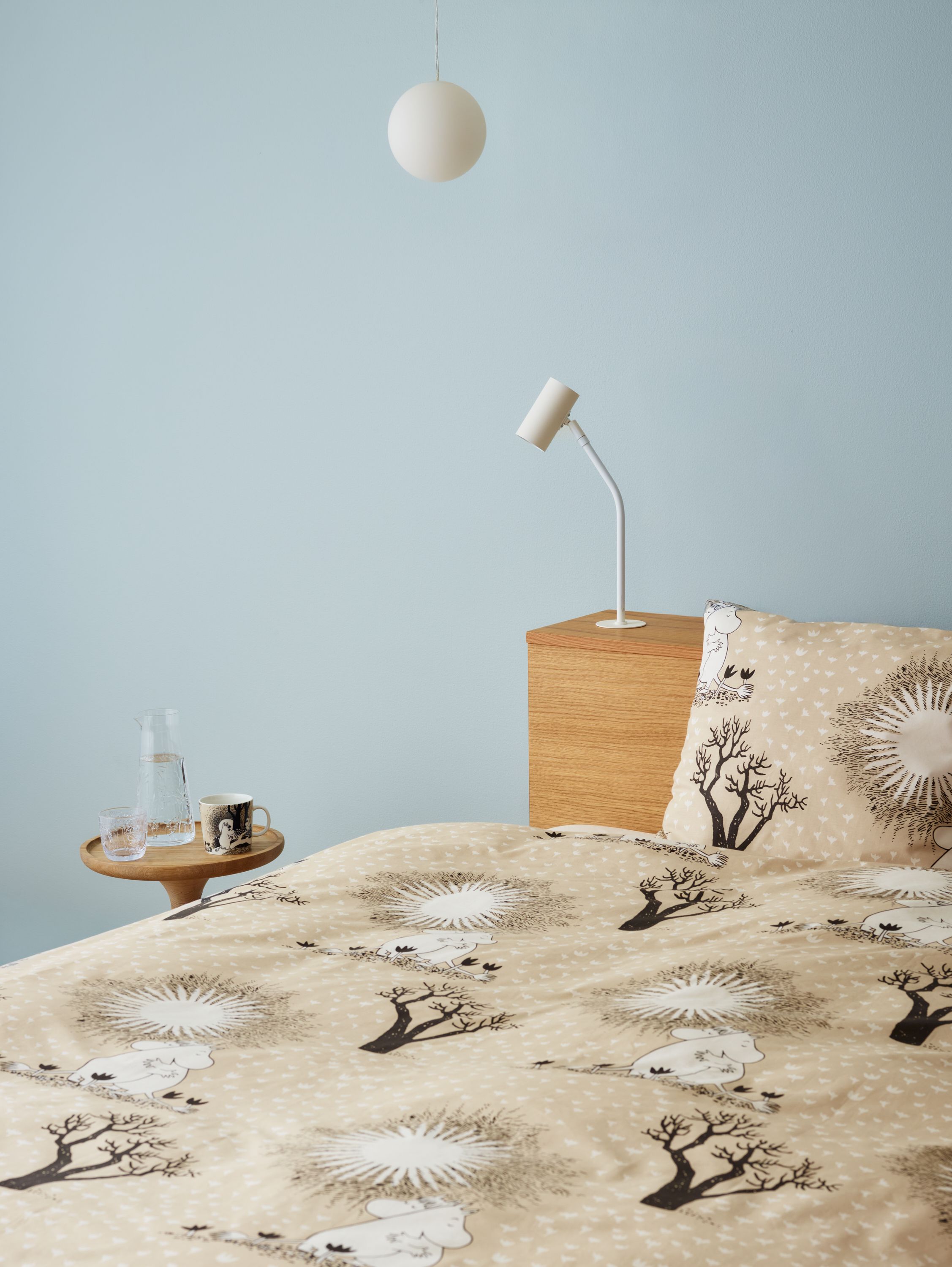 Ljust sovrum med beige Mumin-påslakan, träbord med glas och mugg samt minimalistisk vit lampa mot ljusblå vägg.