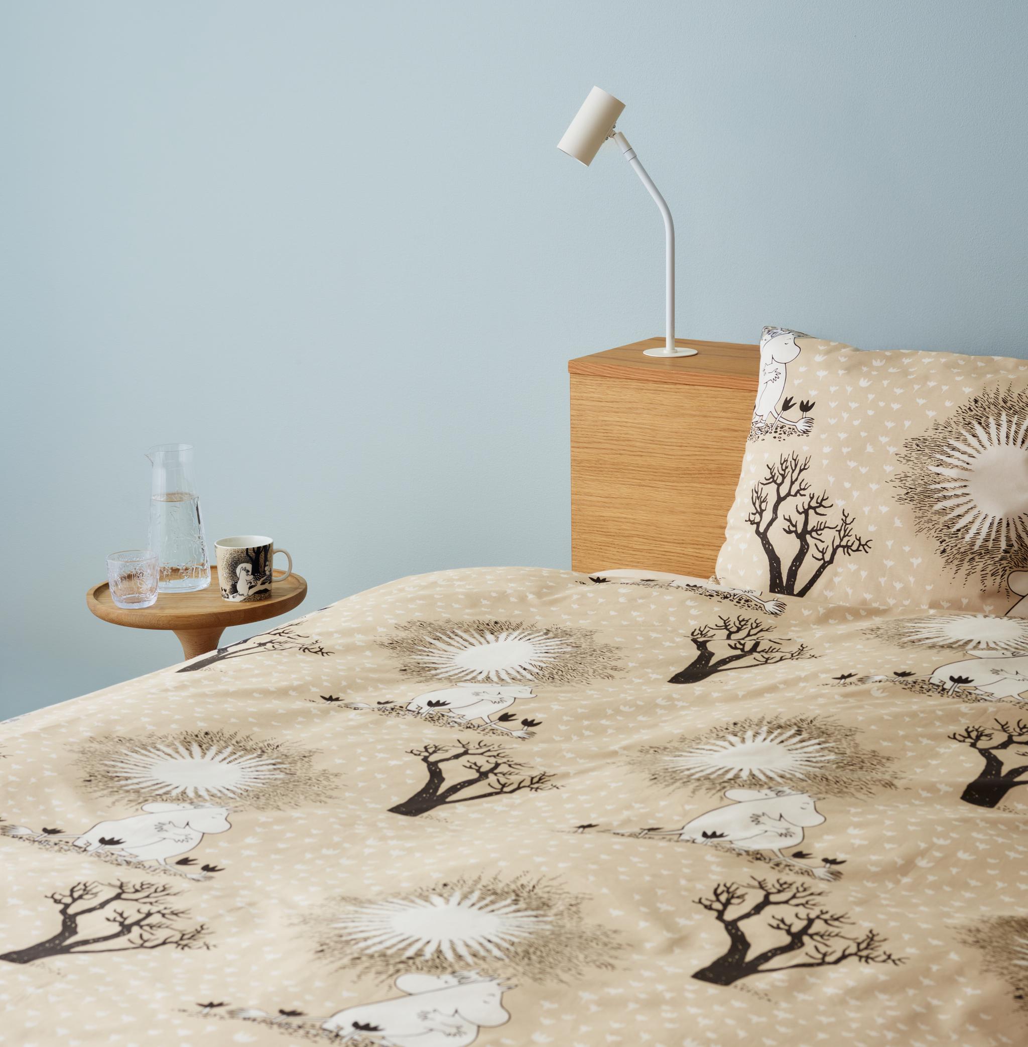 Ljust sovrum med beige Mumin-påslakan, träbord med glas och mugg samt minimalistisk vit lampa mot ljusblå vägg.