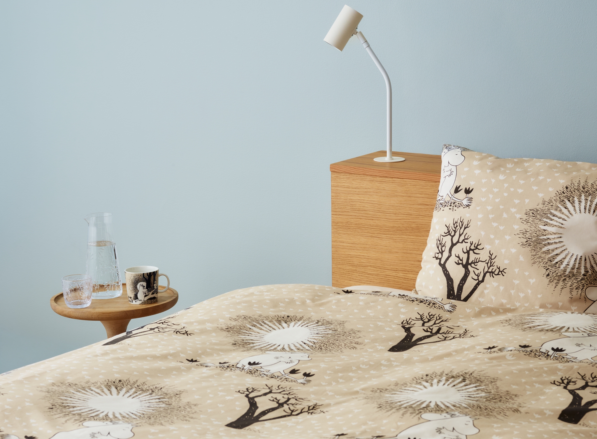 Ljust sovrum med beige Mumin-påslakan, träbord med karaff och mugg samt minimalistisk vit lampa mot ljusblå vägg.