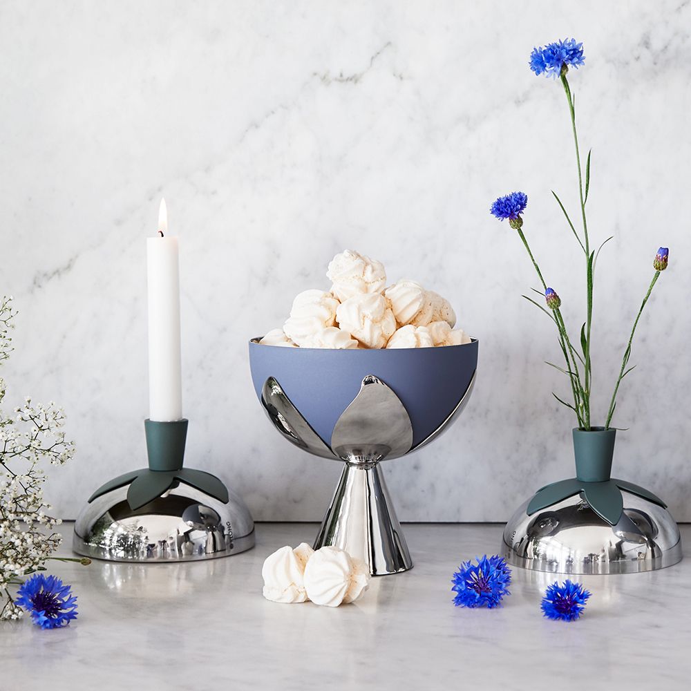 A modern still life with a lit candle, a blue and silver bowl of meringues, and blue cornflowers in a vase, on a marble surface.