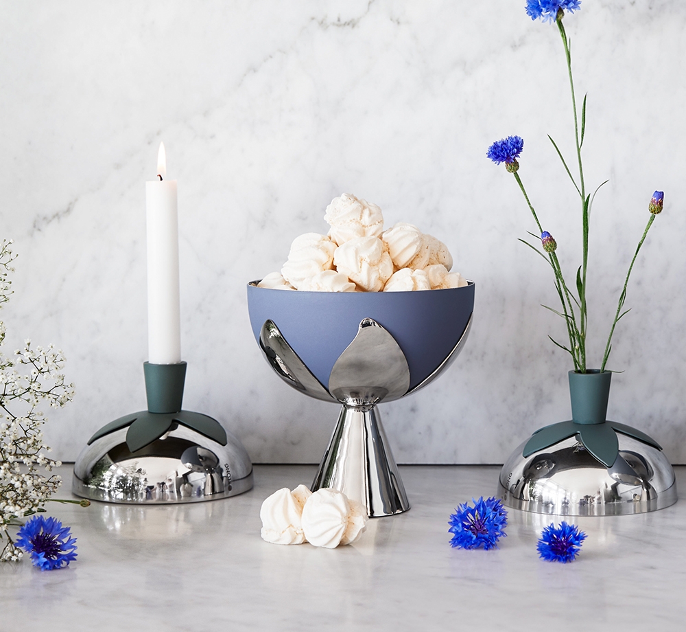 A modern still life with a lit candle, a blue and silver bowl of meringues, and blue cornflowers in a vase, on a marble surface.