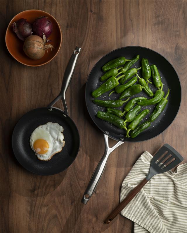 Kaksi paistinpannua puupöydällä, toisessa paistettu muna ja toisessa vihreät paprikat.