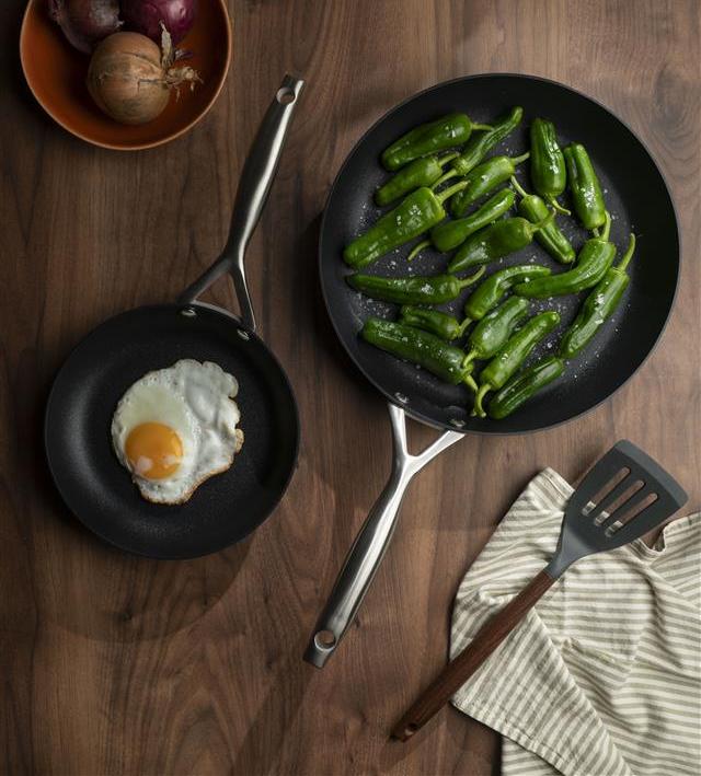 Kaksi paistinpannua puupöydällä, toisessa paistettu muna ja toisessa vihreät paprikat.