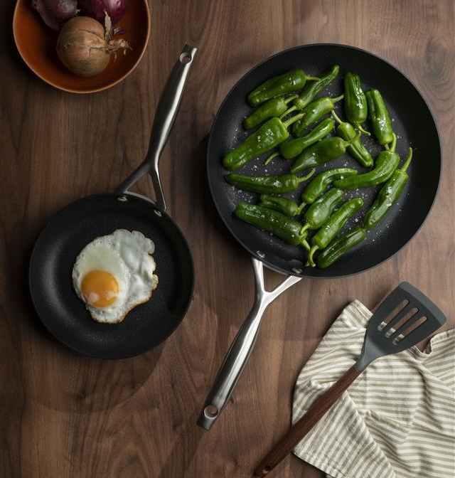 Kaksi paistinpannua puupöydällä, toisessa paistettu muna ja toisessa vihreät paprikat.