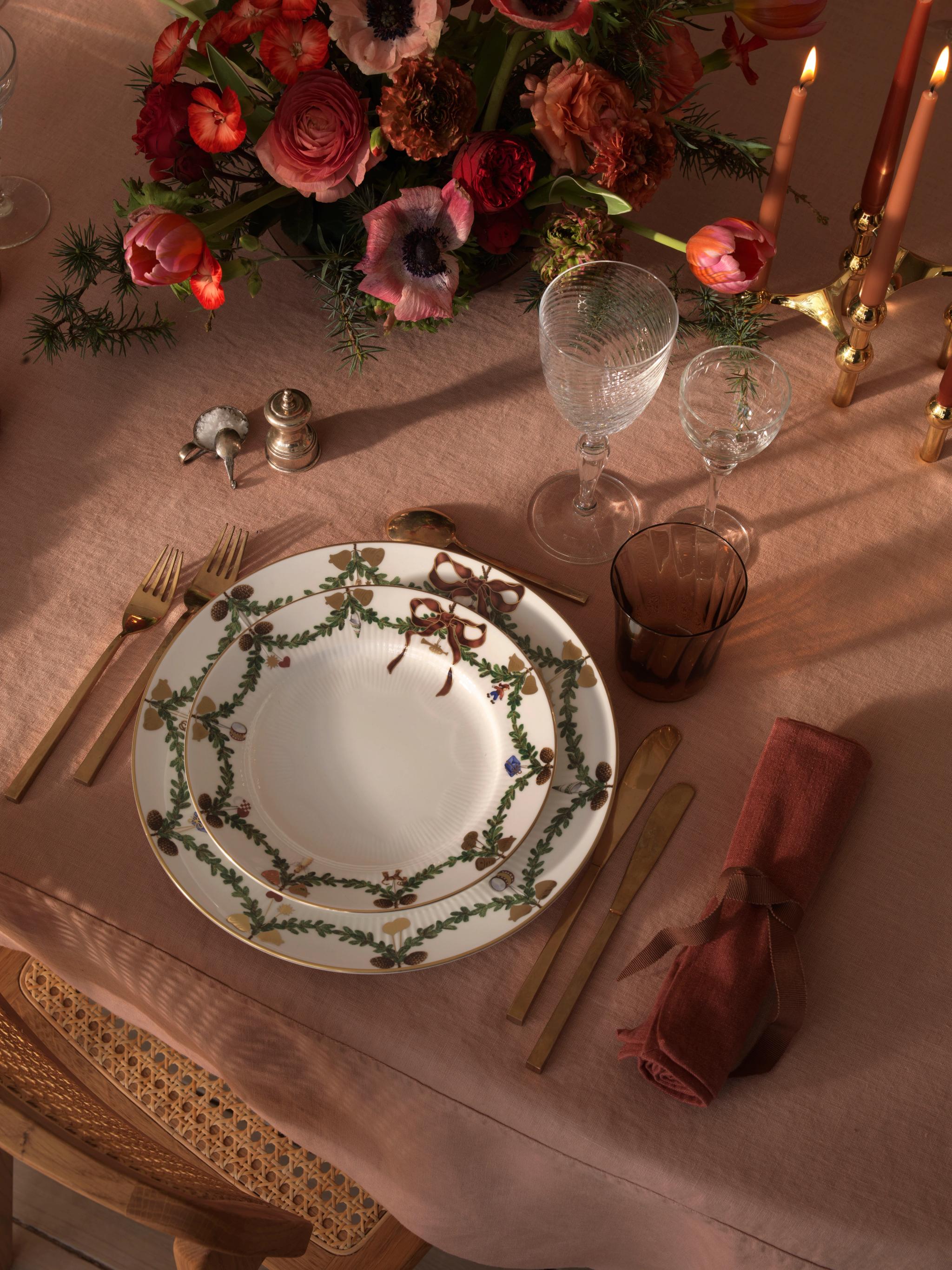 En festlig dukning med vita tallrikar med grön girlangdesign, gyllene bestick, rosa servetter, glas, en blommig mittdekoration och tända ljus på en rosa duk.
