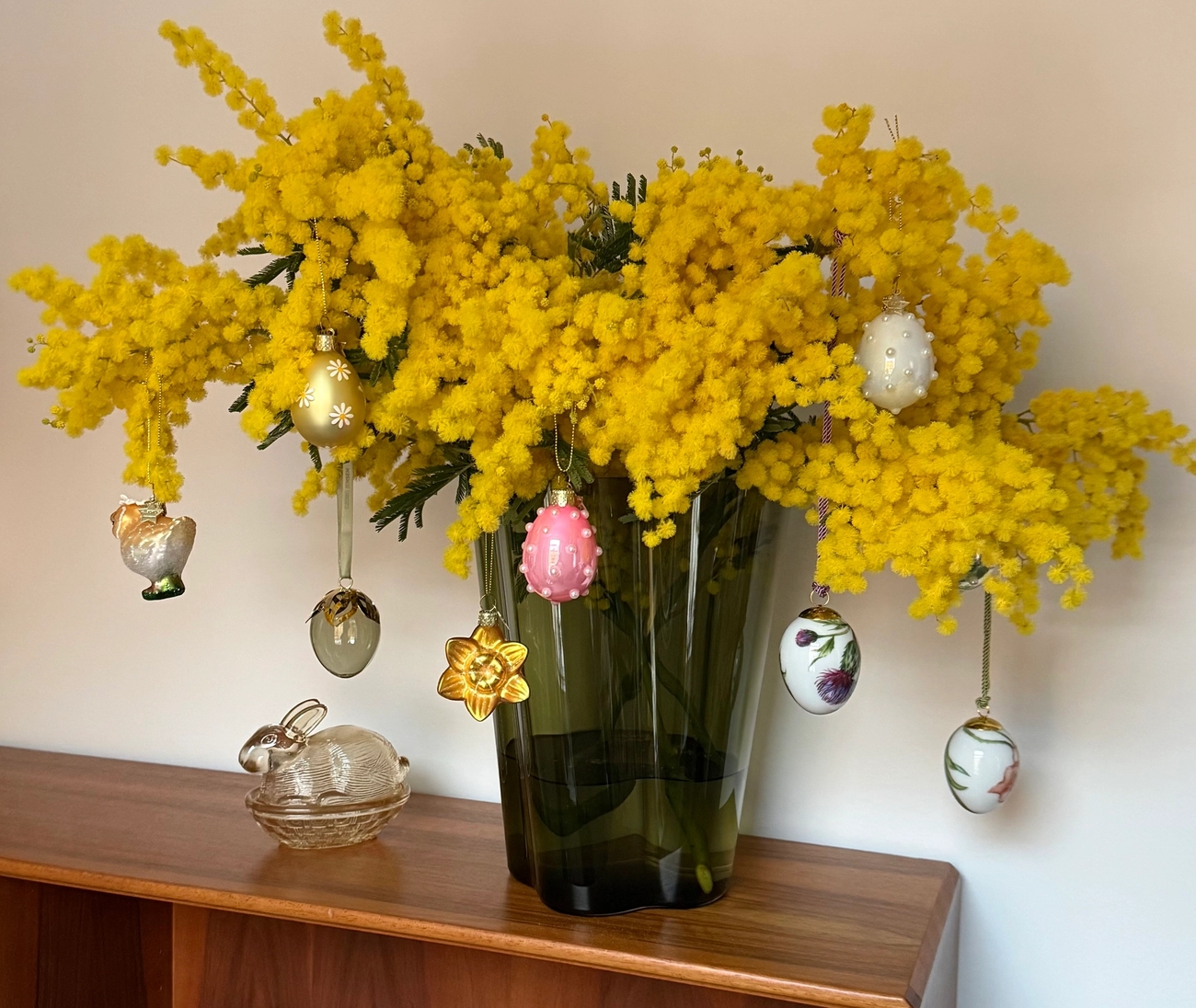 En bukett med knallgule mimosablomster i en vase, pyntet med hengende påskeegg
