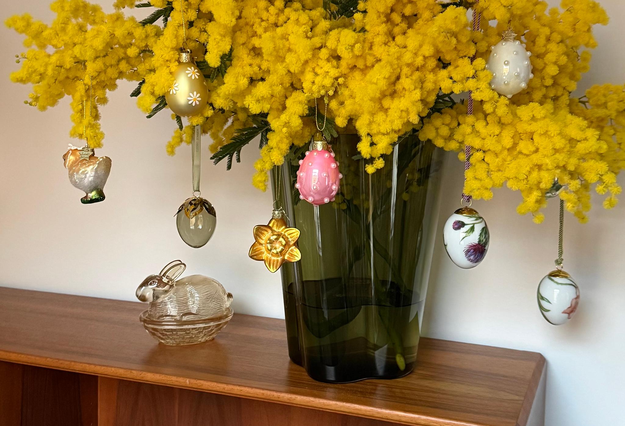 Pääsiäiskoristein koristeltu maljakko, jossa on keltaisia mimoosakukkia, lasisen kanihahmon vieressä puisella hyllyllä.