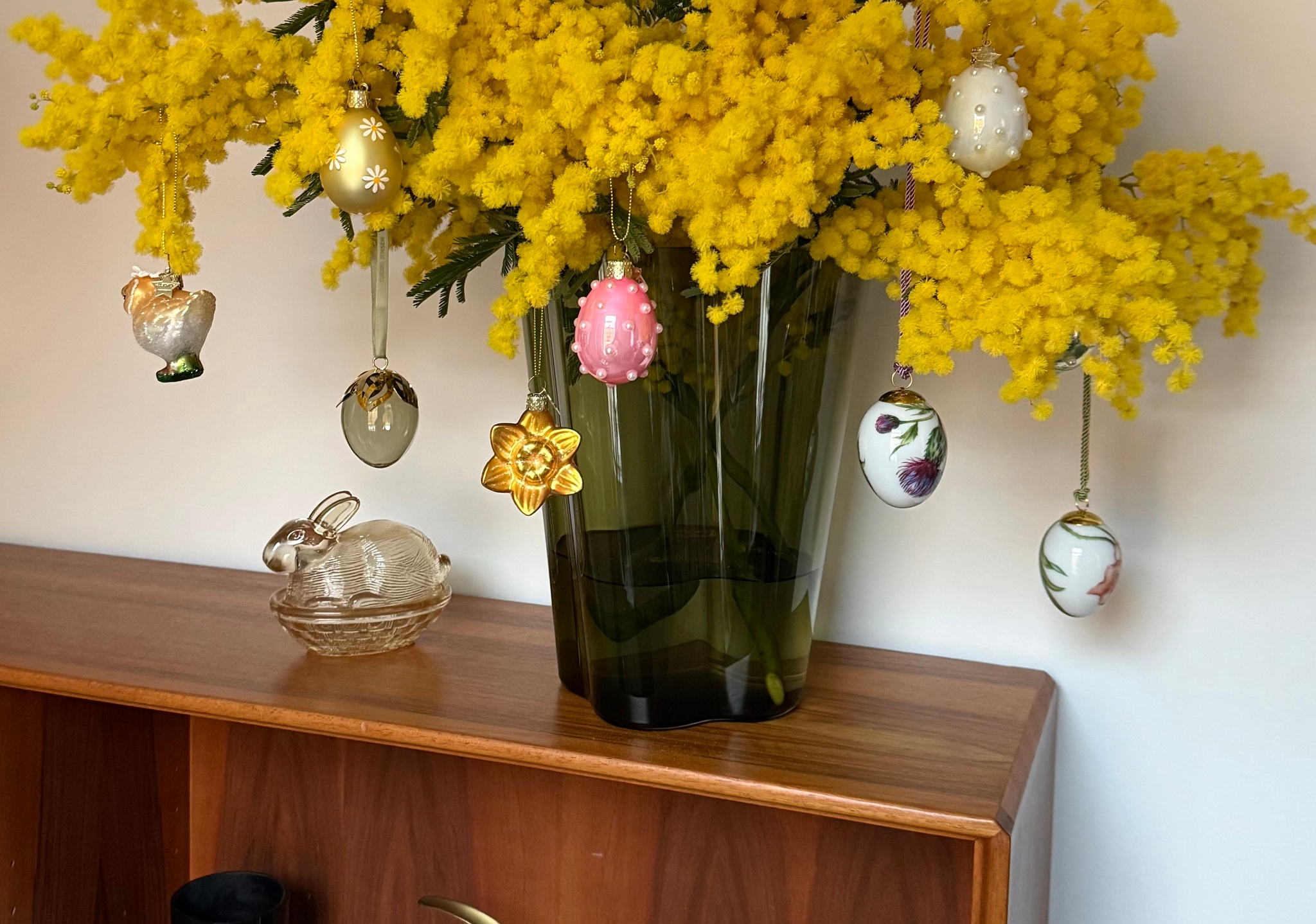 Pääsiäiskoristein koristeltu maljakko, jossa on keltaisia mimoosakukkia, lasisen kanihahmon vieressä puisella hyllyllä.