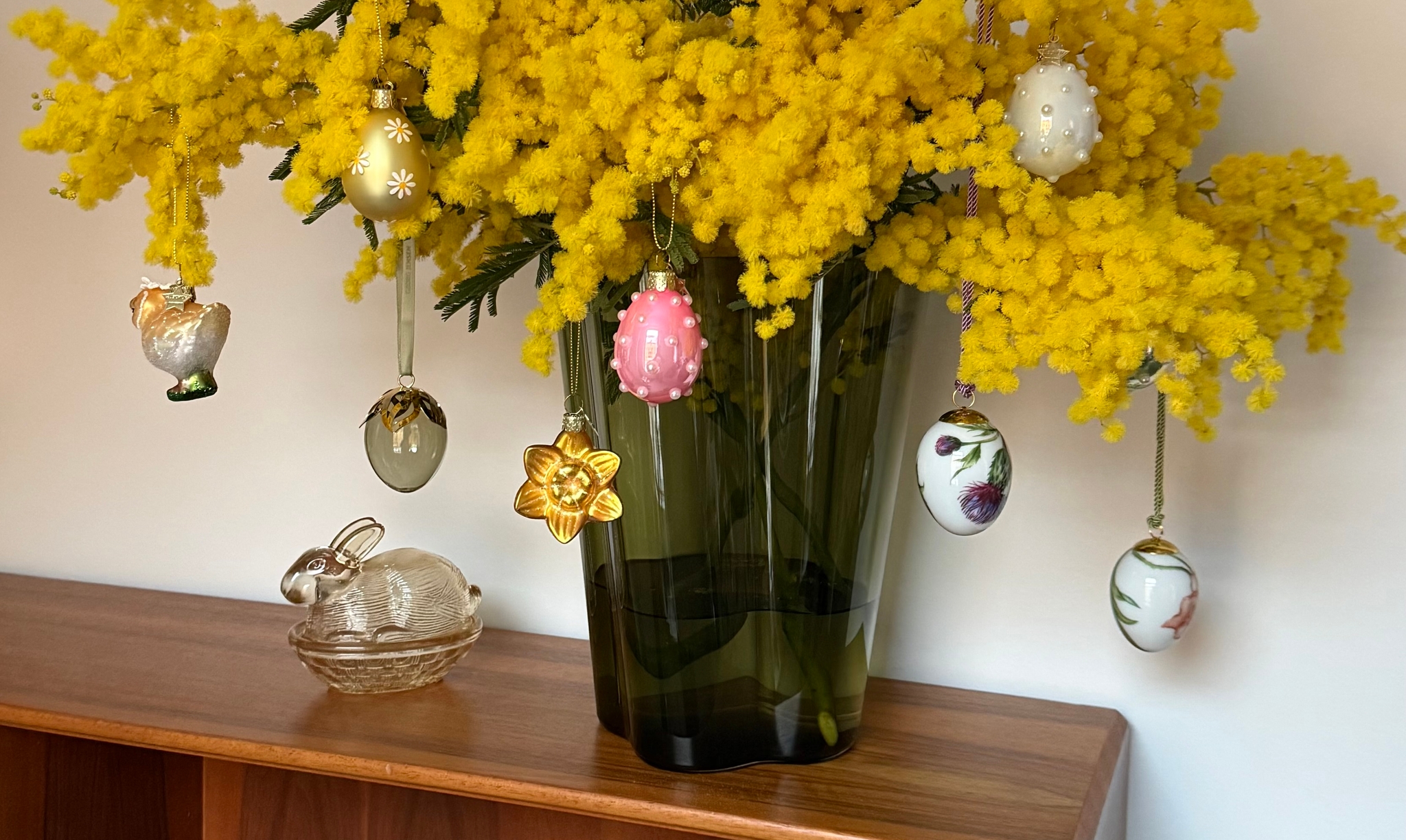 Keltaisia mimosakukkia maljakossa, jota koristavat pääsiäiskoristeet, lasikanin vieressä puupinnalla.