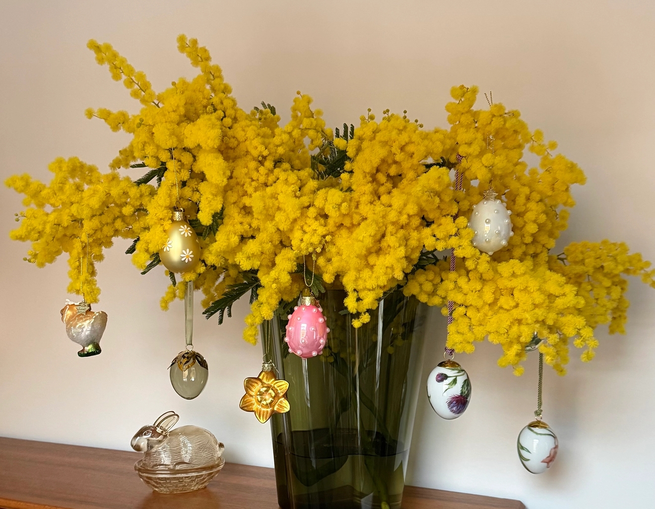 En bukett med knallgule mimosablomster i en vase, pyntet med hengende påskeegg
