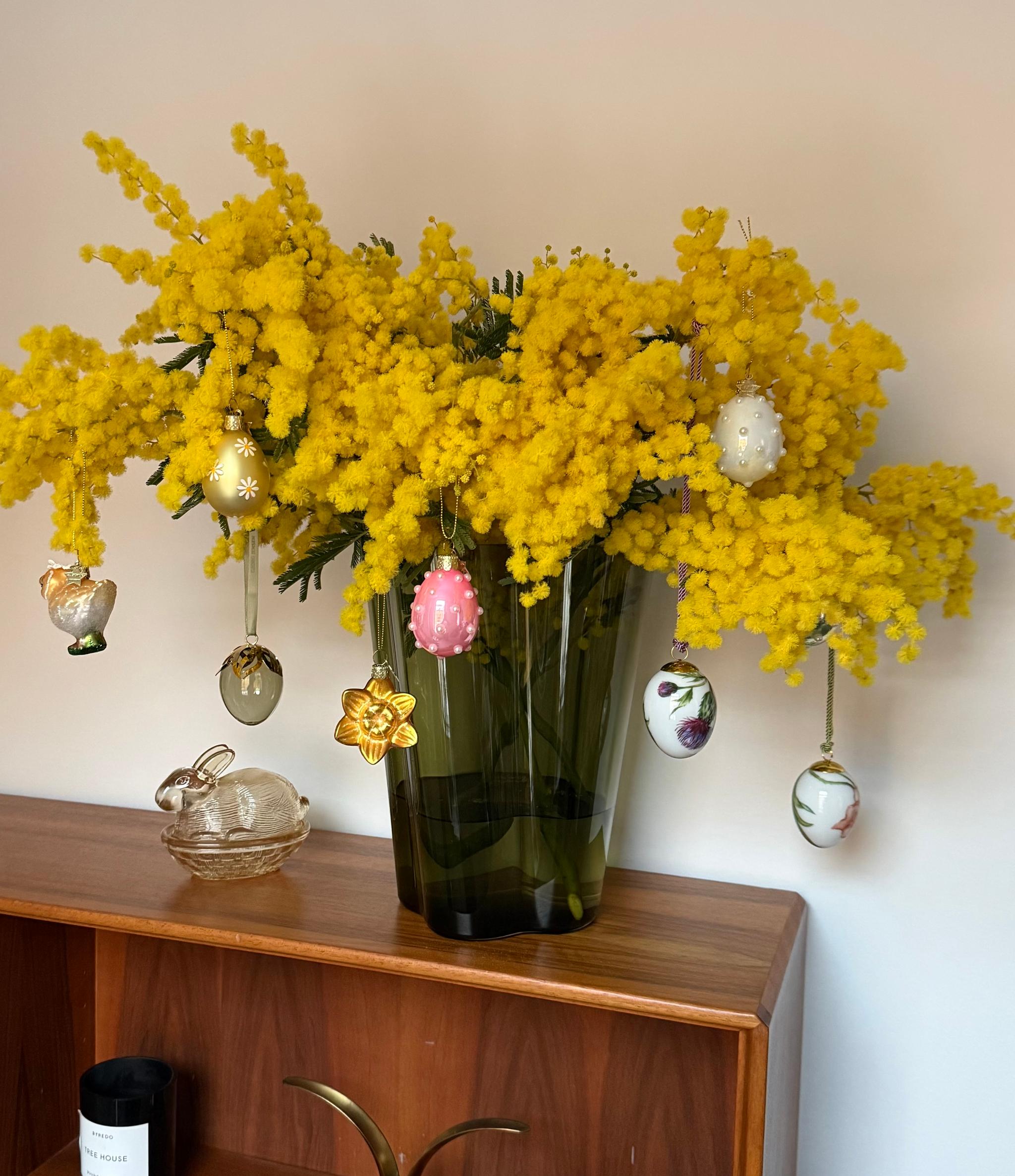 En bukett med klargula mimosablommor i en grön vas, dekorerad med hängande påskägg och en påsklilja, bredvid en glasfigur föreställande en kanin på en träyta.